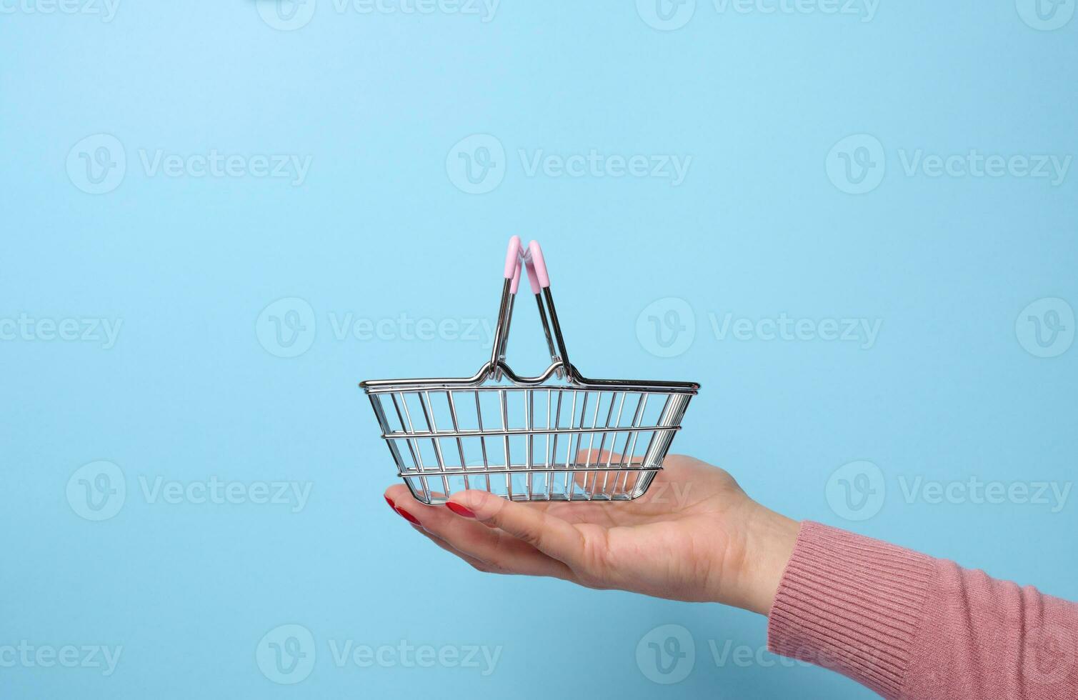 mulher mão segurando uma miniatura metal compras cesta em uma azul fundo foto