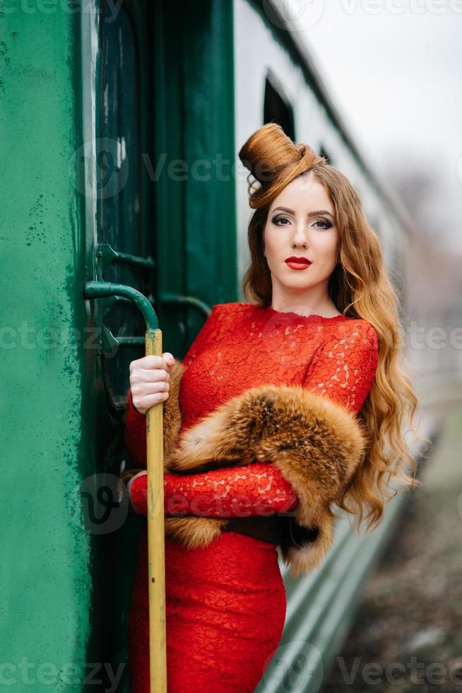 jovem com cabelo ruivo em um vestido vermelho brilhante perto de um velho carro de passageiros foto