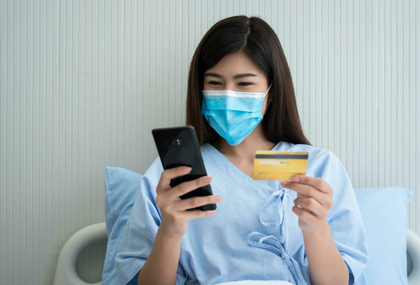 feliz ásia mulher vestindo uma médico mascarar e segurando zombar acima crédito seguro cartão e Smartphone dentro uma hospital cama para Verifica saúde seguro elegibilidade. seguro política de banco, Forma de pagamento médico foto