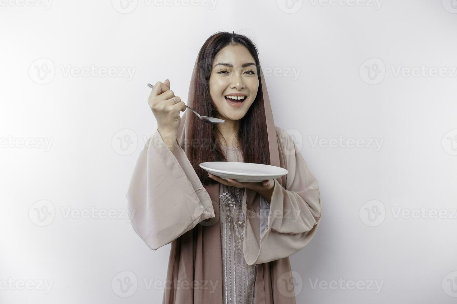 uma sorridente ásia muçulmano mulher é jejum e com fome e segurando e apontando para uma prato foto