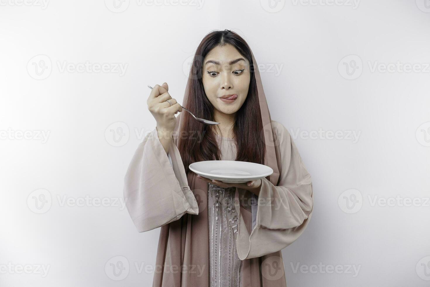 uma sorridente ásia muçulmano mulher é jejum e com fome e segurando e apontando para uma prato foto