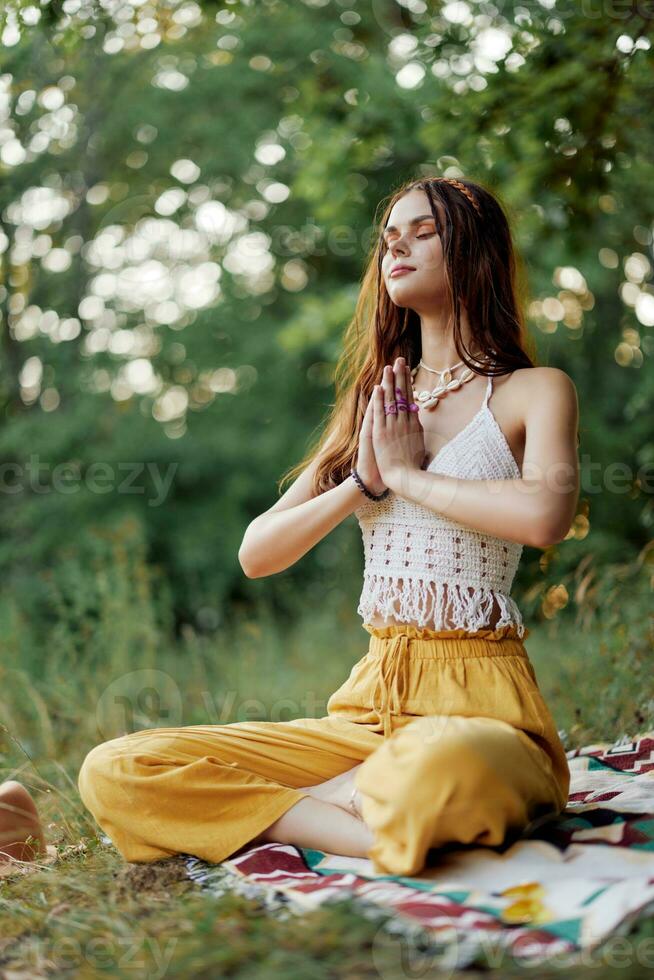 uma jovem hippie mulher medita dentro natureza dentro a parque, sentado dentro uma lótus posição em dela colorida xadrez e desfrutando harmonia com a mundo dentro roupas ecológicas foto