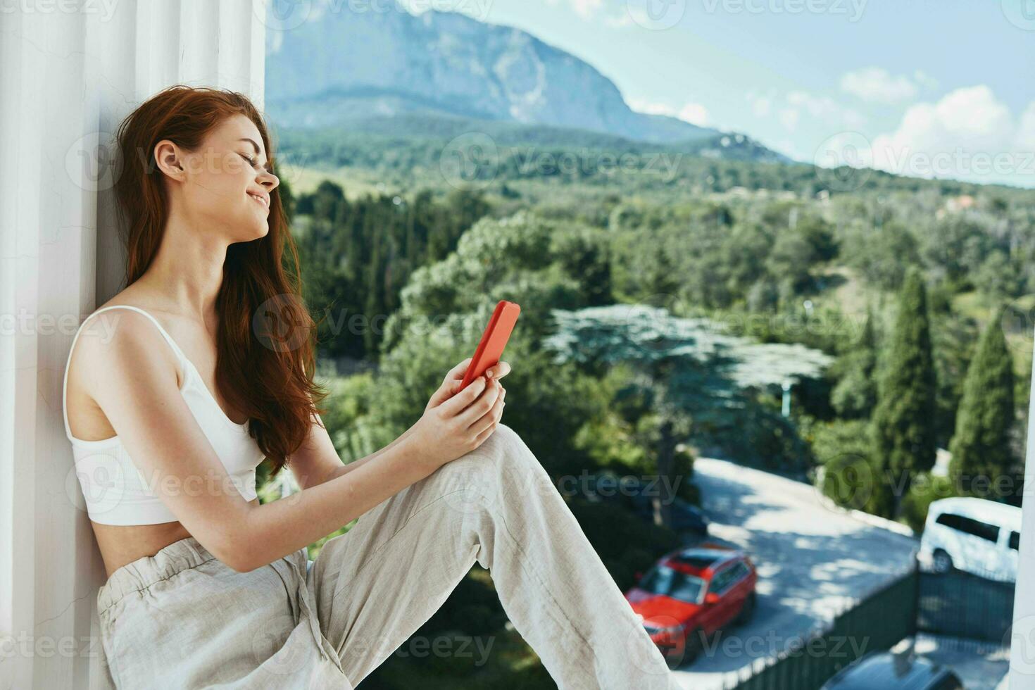 atraente jovem mulher sentado em a sacada com telefone lindo montanha Visão verão inalterado foto