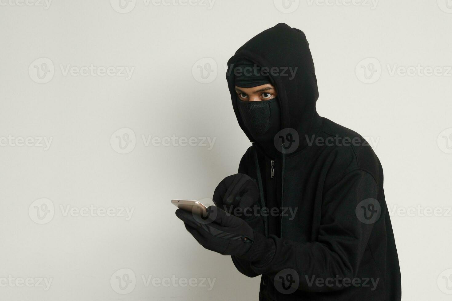 retrato do misterioso homem vestindo Preto moletom com capuz e mascarar fazendo hacking atividade em Móvel telefone, hacker segurando uma Smartphone. isolado imagem em cinzento fundo foto