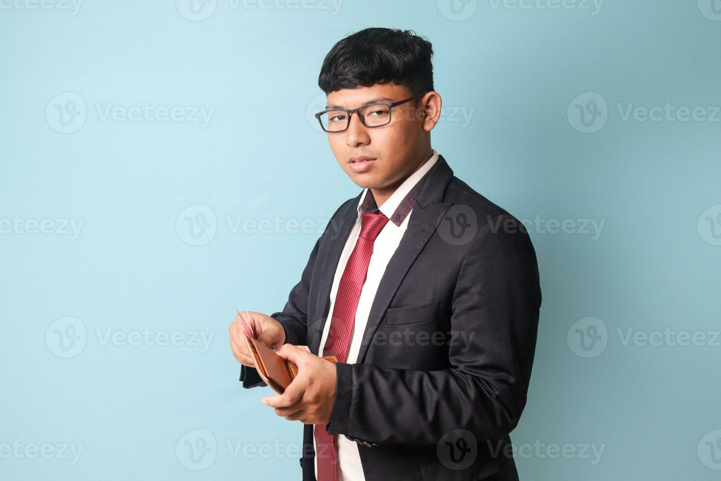 retrato do jovem ásia o negócio homem dentro casual terno segurando couro carteira com sério expressões. isolado imagem em azul fundo foto