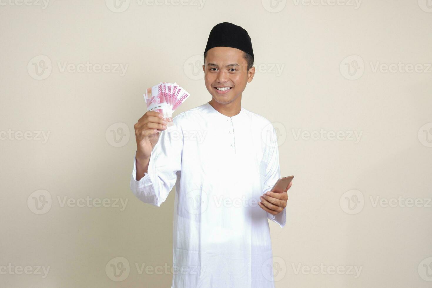 retrato do atraente ásia muçulmano homem dentro branco camisa mostrando 1 cem mil rupia enquanto usando Móvel telefone. financeiro e poupança conceito. isolado imagem em cinzento fundo foto