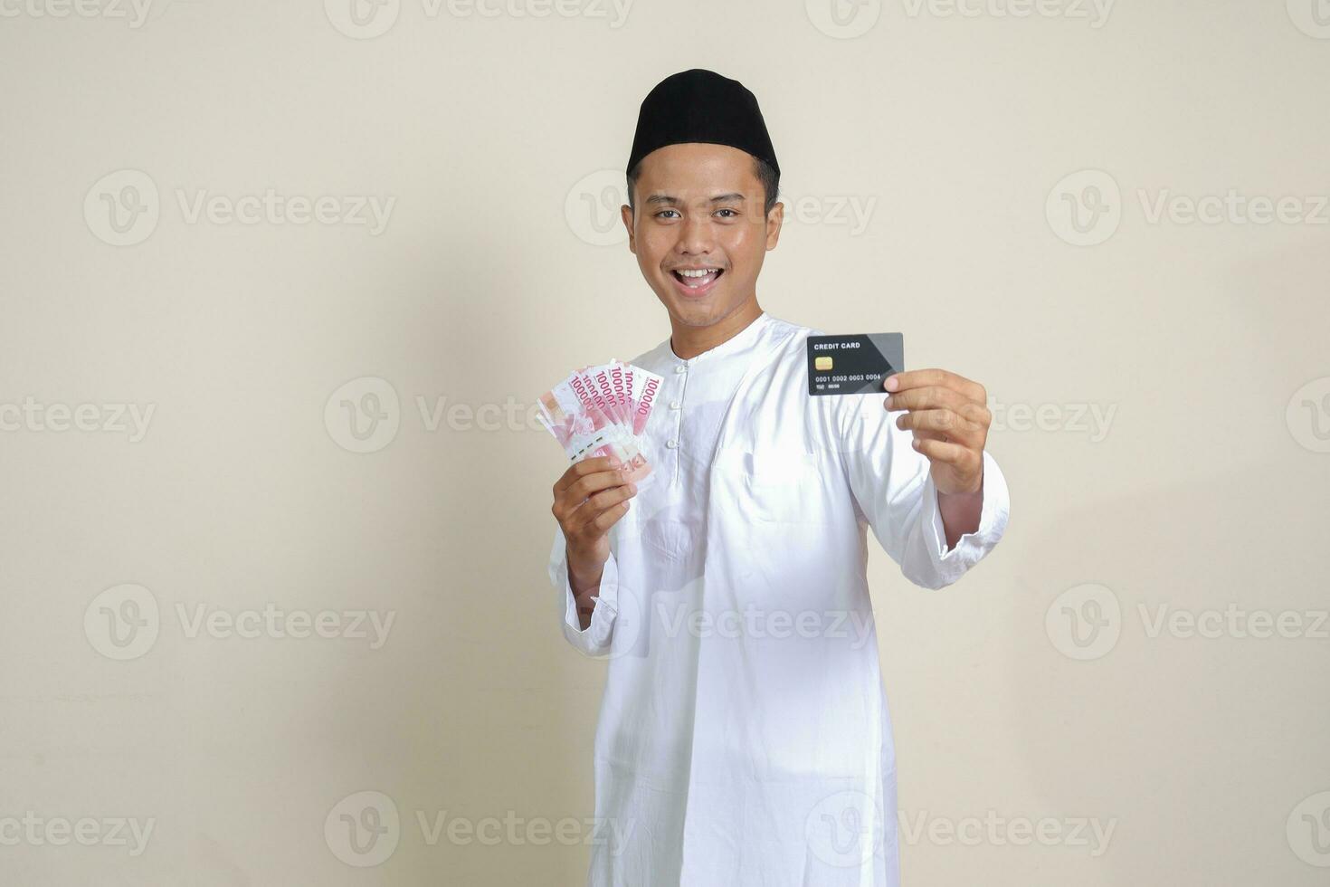 retrato do atraente ásia muçulmano homem dentro branco camisa com calota craniana segurando 1 cem mil rupia e apresentando crédito cartão. isolado imagem em cinzento fundo foto