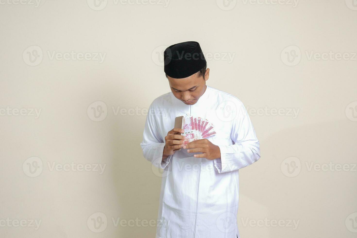 retrato do atraente ásia muçulmano homem dentro branco camisa mostrando 1 cem mil rupia enquanto usando Móvel telefone. financeiro e poupança conceito. isolado imagem em cinzento fundo foto