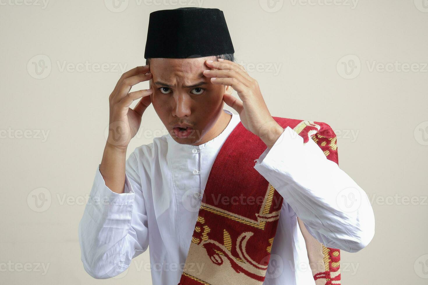 retrato do atraente ásia muçulmano homem dentro branco camisa com calota craniana sentindo-me bonito e confiante. tocante dele templos para faço uma estilo. isolado imagem em cinzento fundo foto