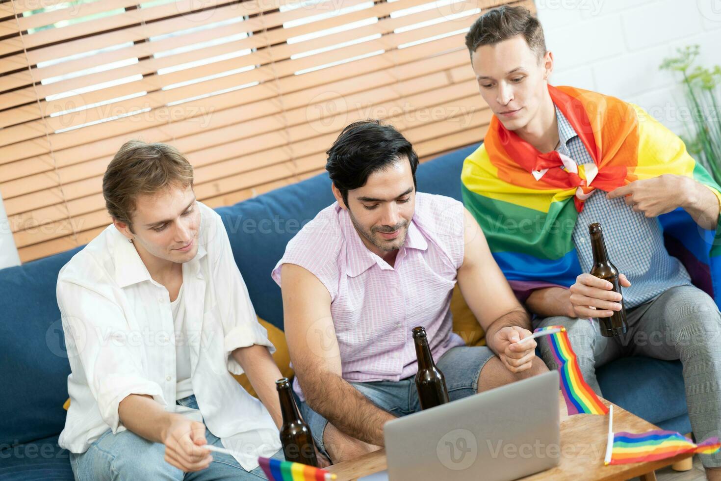 triplo jovem homem sentado dentro a vivo quarto enquanto usando computador portátil para vídeo ligar e segurando e acenando arco Iris lgbt orgulho bandeira. lgbtq pessoas estilo de vida. celebrando a orgulho dia. foto
