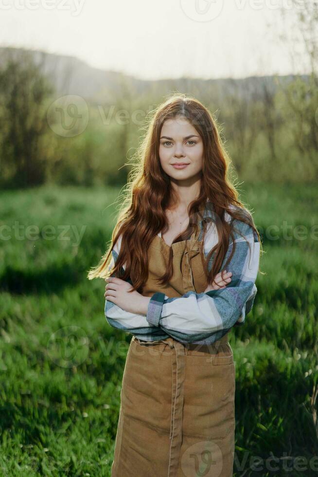 uma agricultor mulher dentro dela trabalhos roupas, xadrez camisa e avental, carrinhos dentro a campo em a verde Relva e sorrisos dentro a configuração Sol foto