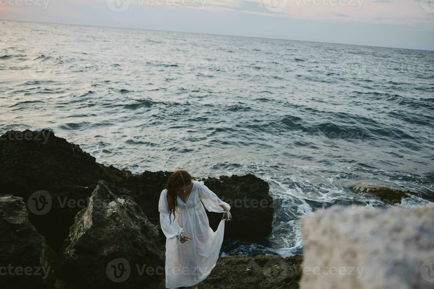 uma mulher dentro uma branco vestir carrinhos descalço em a pedras de a oceano foto