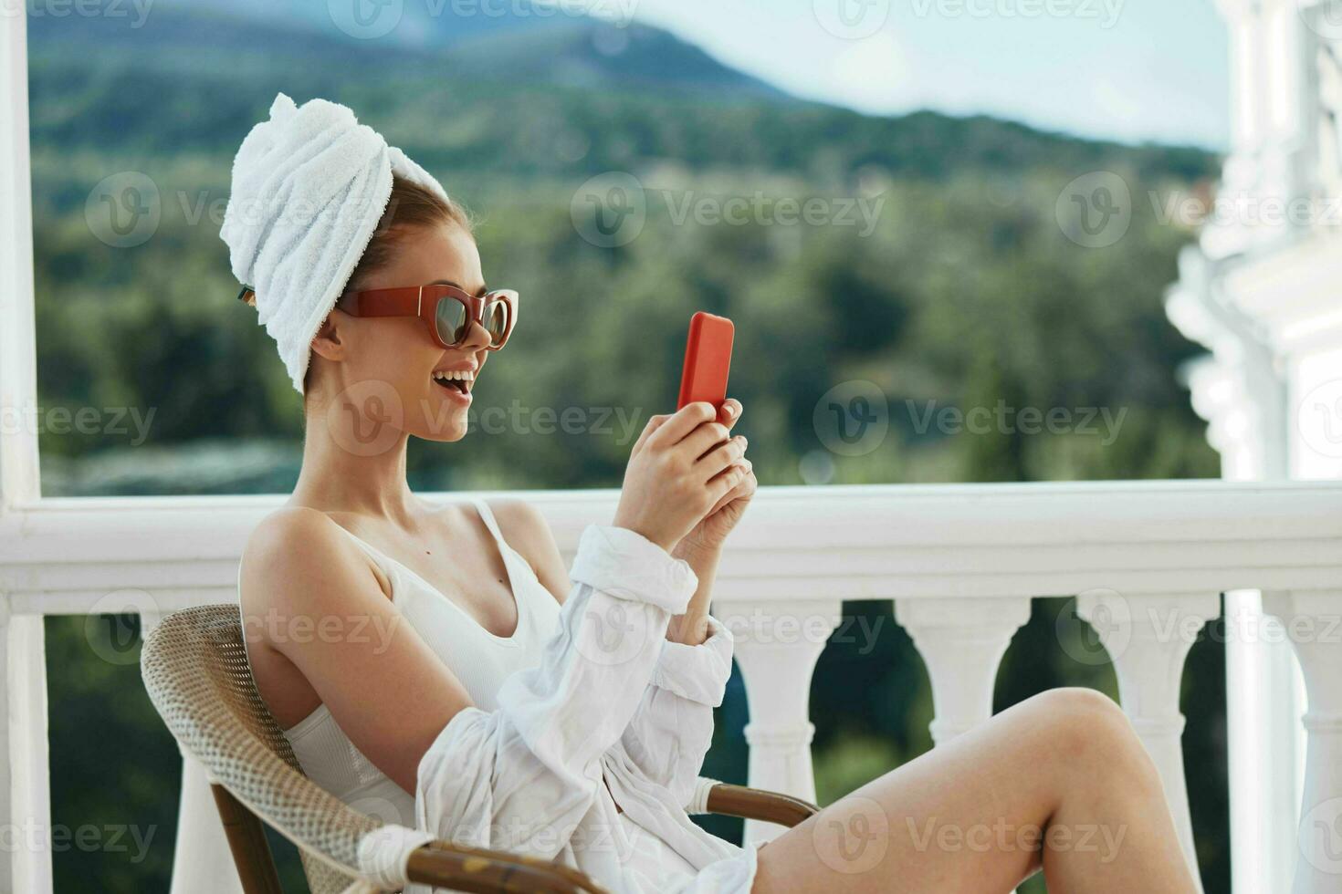 lindo mulher dentro uma branco camisa senta em a sacada com uma telefone preguiçoso manhã foto