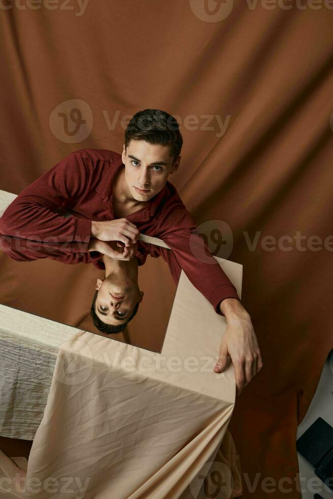 uma homem com uma espelho senta às uma mesa em uma tecido fundo e uma Castanho camisa foto