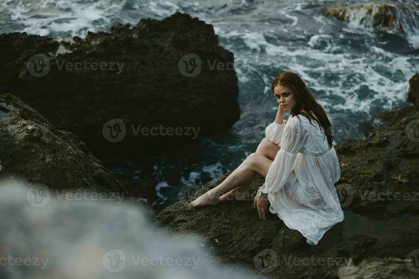 uma mulher dentro uma branco Casamento vestir é sentado descalço em uma onda penhasco foto