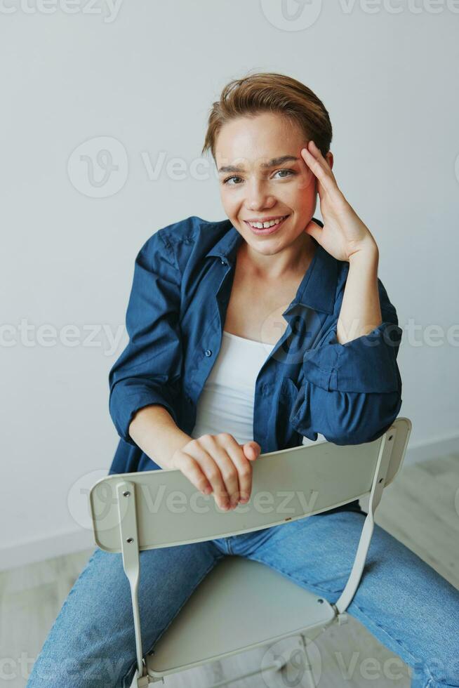 uma jovem mulher sentado dentro uma cadeira às casa sorridente com dentes com uma curto corte de cabelo dentro jeans e uma jeans camisa em uma branco fundo. menina natural poses com não filtros foto