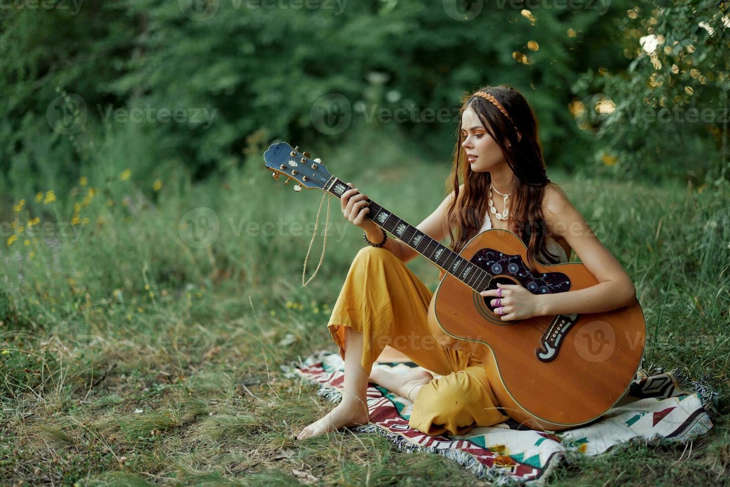 jovem hippie mulher sentado ao ar livre em uma xadrez e jogando dela guitarra em a margem do rio para dentro a pôr do sol foto