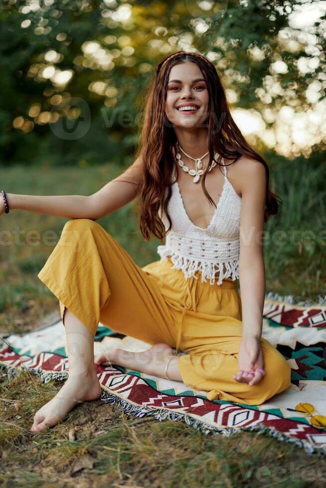 uma jovem lindo hippie mulher senta em a terra dentro a outono dentro eco roupas dentro a pôr do sol luz e sorrisos Atenciosamente para a Câmera foto