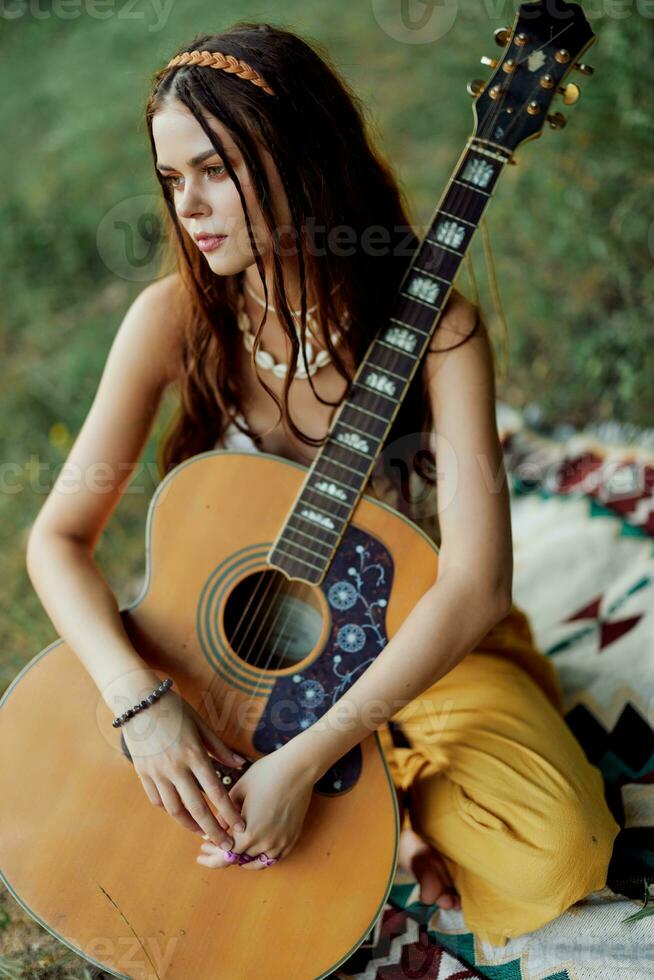 uma menina dentro roupas ecológicas hippie sentado com uma guitarra e olhando às uma pôr do sol dentro a verão foto