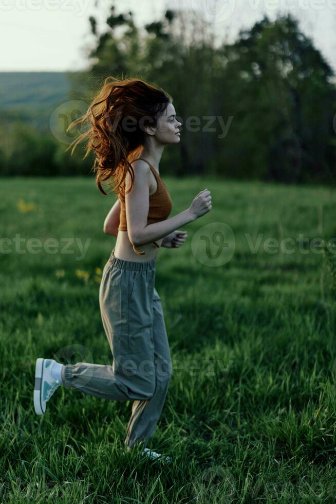 uma mulher com grandes vermelho cabelo trabalho Fora e corre em a verde Relva dentro a parque dentro moletom e tênis foto