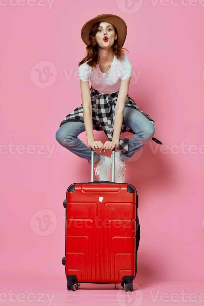 alegre mulher turista senta em uma vermelho mala de viagem voar aeroporto foto