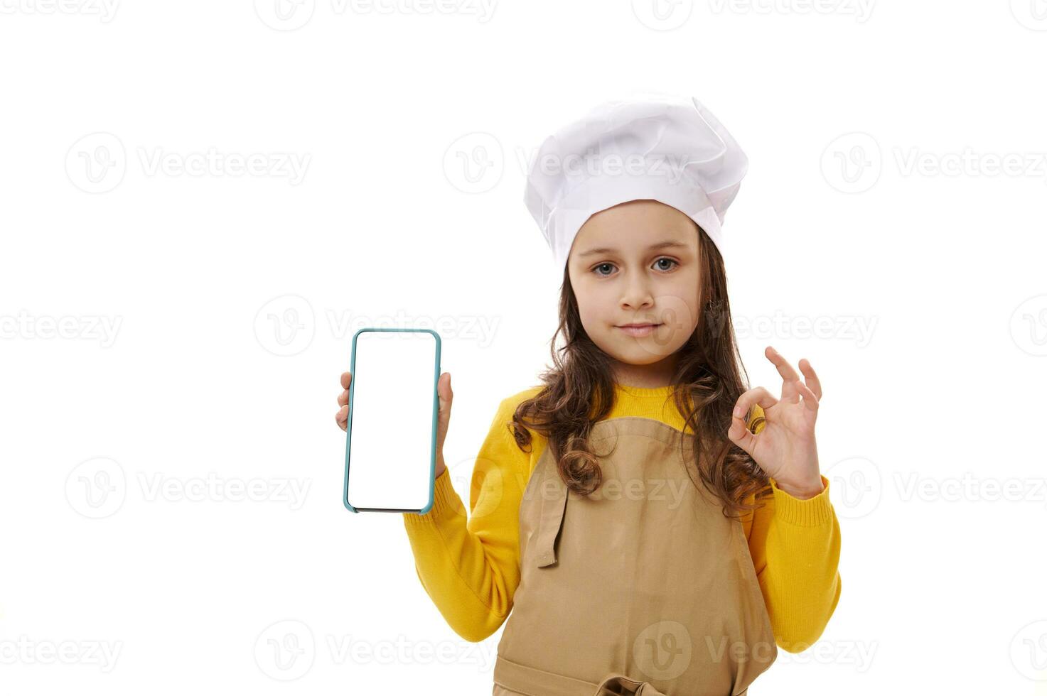 pequeno garota, chefe de cozinha pasteleiro, gestos com Está bem placa e mostra às Câmera uma Smartphone com em branco branco digital tela foto