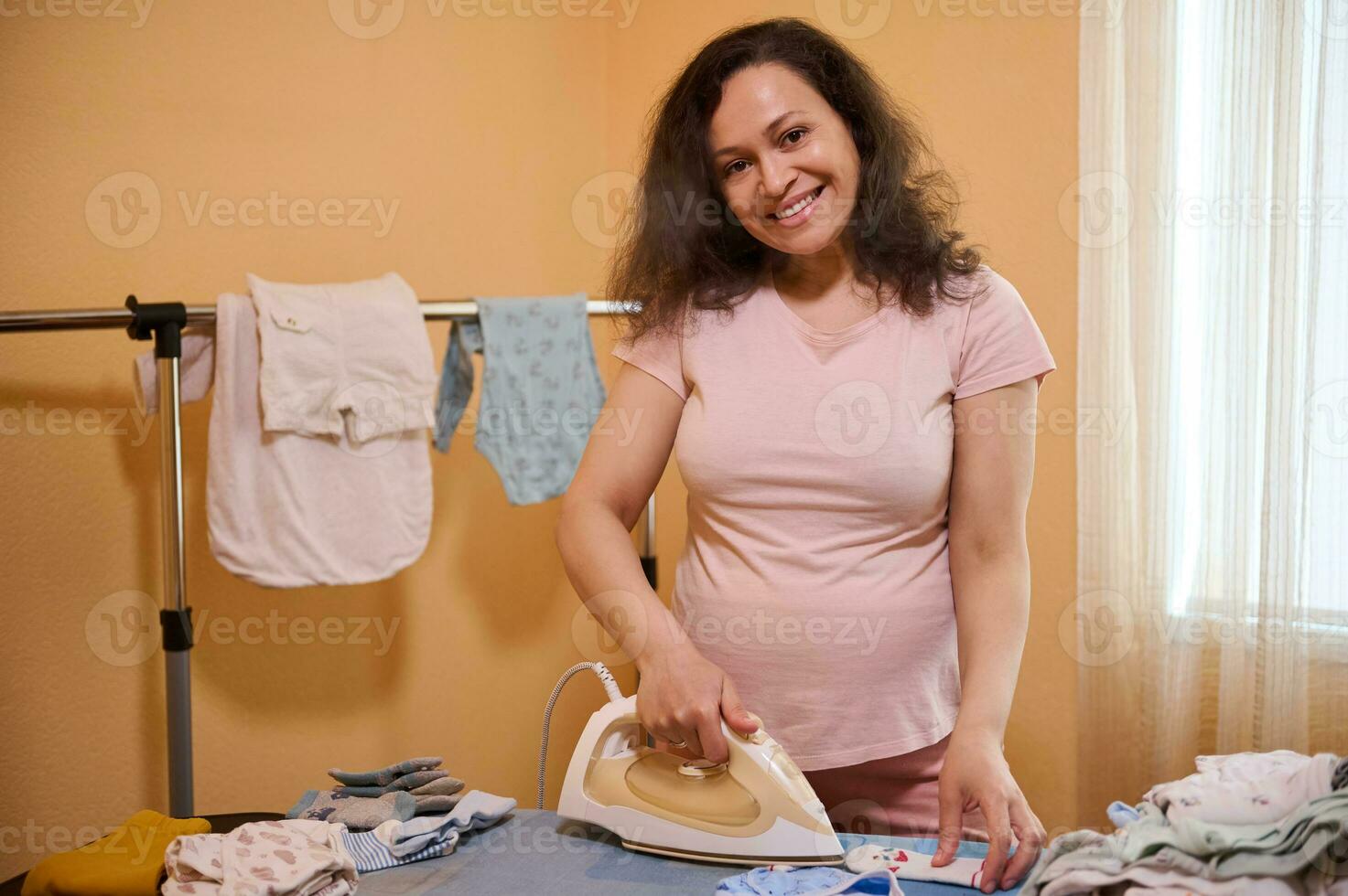 meio envelhecido multi étnico positivo sorridente grávida mulher, futuro mãe passar roupa bebê roupas, preparando saco para criança nascimento foto