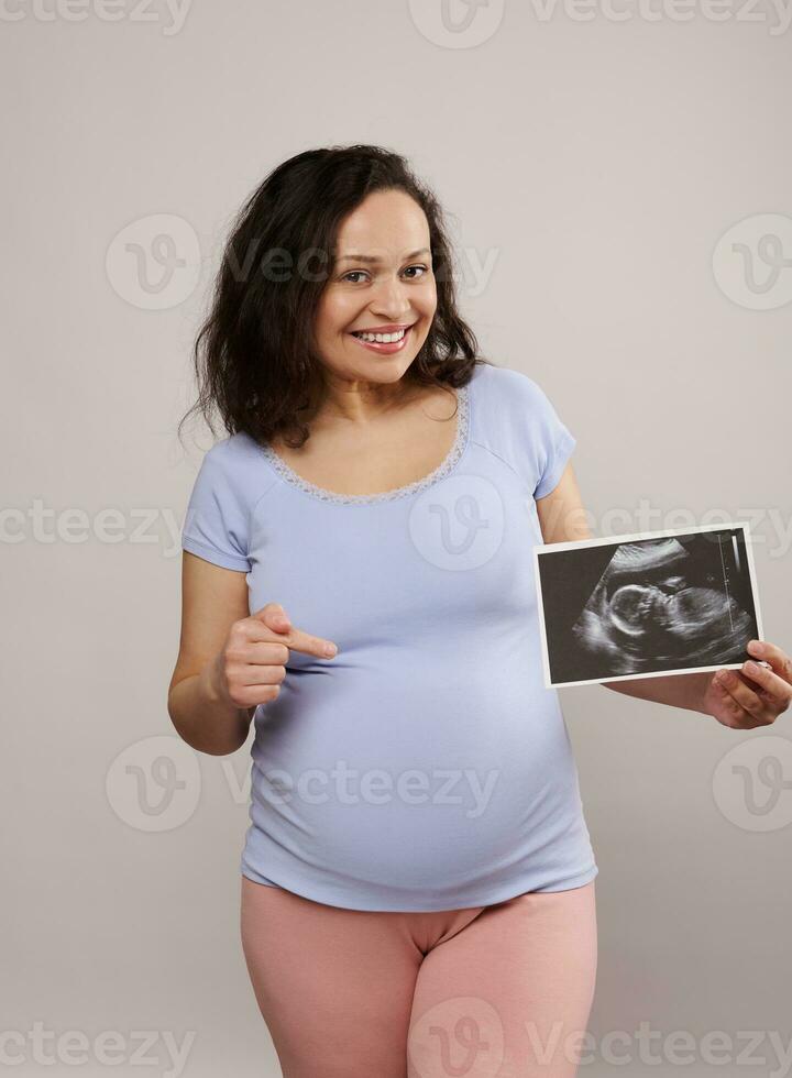 sorridente grávida mulher mostrando dela recém-nascido bebê sonografia para a Câmera e apontando às dela barriga, isolado em branco foto