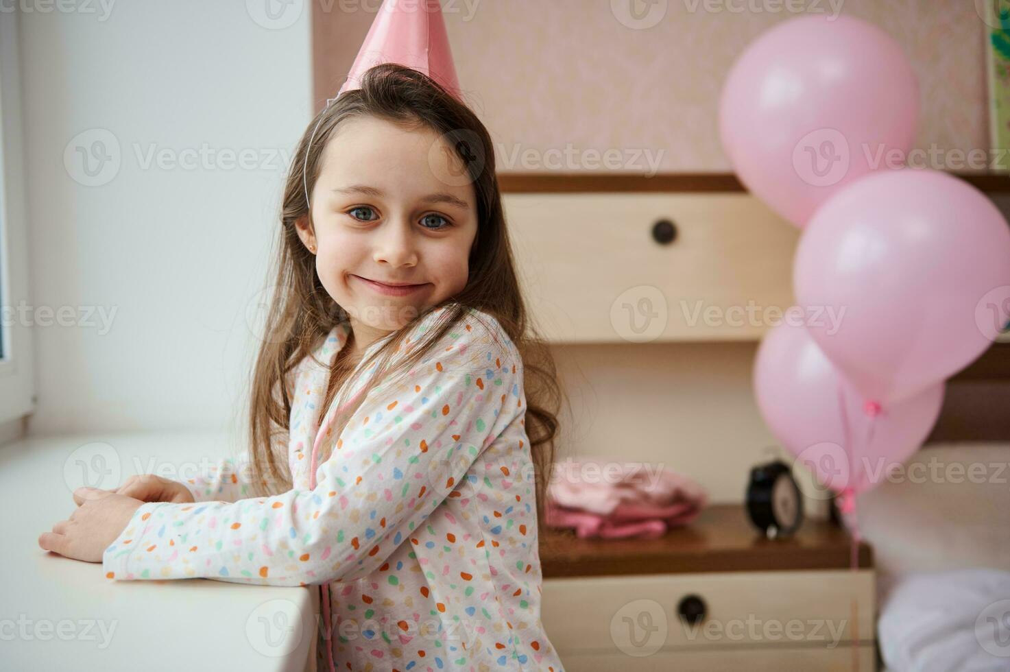 fofa aniversário menina sorrisos olhando para dentro Câmera, em pé de janela dentro dela quarto decorado com Rosa inflável balões foto