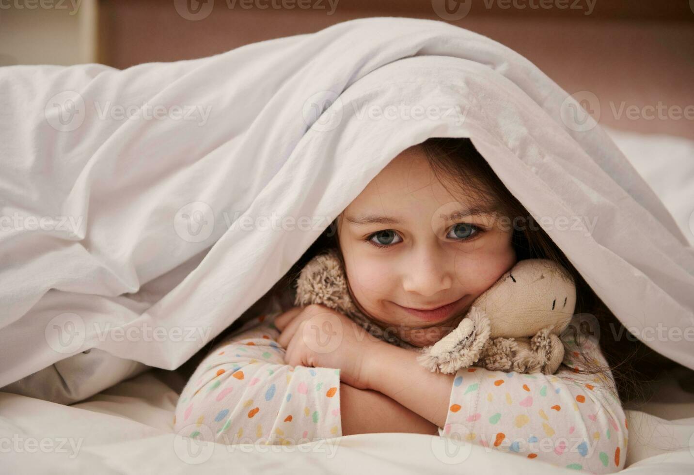 adorável criança menina colheita Fora a partir de suave cobertor, fofinho sorridente olhando às Câmera, suavemente abraçando dela fofa pelúcia brinquedo foto