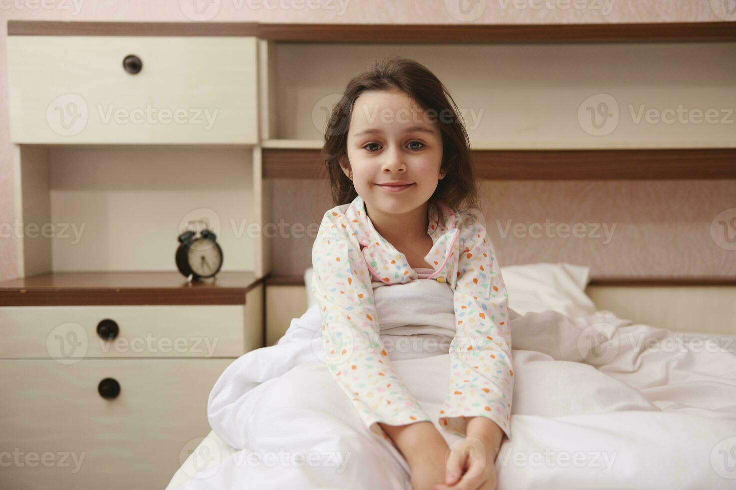 adorável pequeno criança menina dentro pijamas, sorridente olhando às Câmera, sentado dentro cama às dela acolhedor quarto. feliz crianças foto