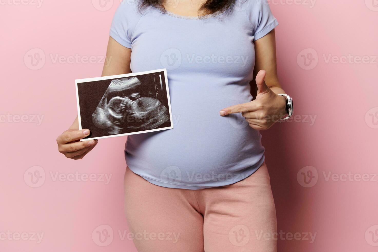 meio do grávida mulher mostrando dela recém-nascido bebê sonografia e apontando às dela barriga, isolado em Rosa fundo foto