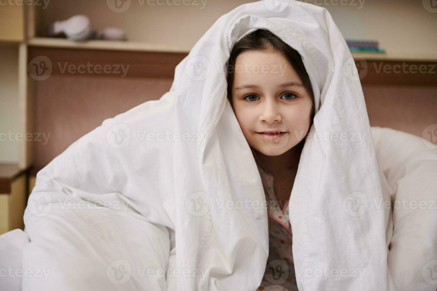 adorável caucasiano pequeno criança menina se escondendo dentro branco suave cobertor, olhando às Câmera, fofinho sorridente olhando às Câmera foto