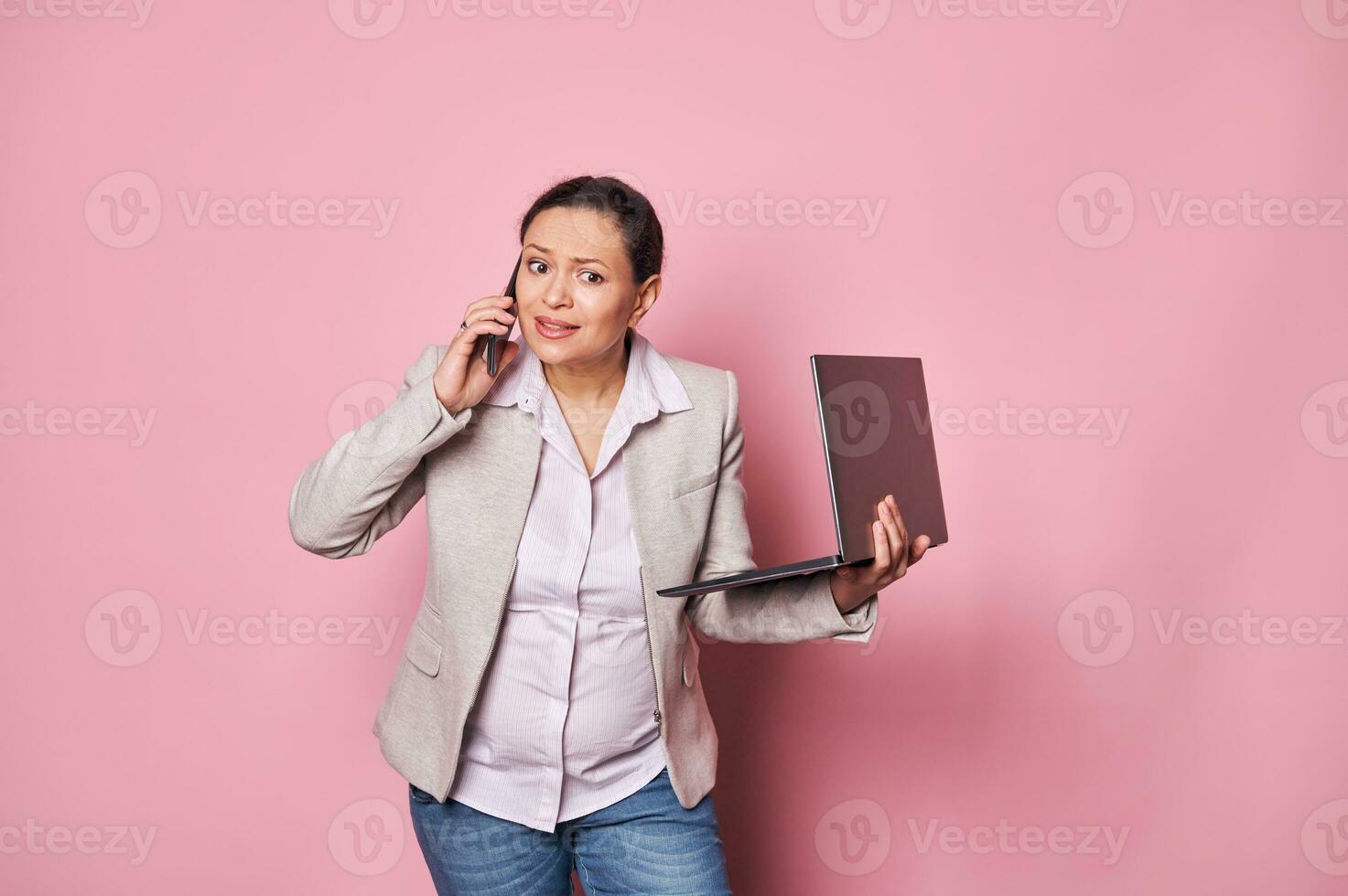 grávida o negócio mulher, segurando computador portátil, falando em Móvel telefone, expressando desapontamento, isolado Rosa fundo foto
