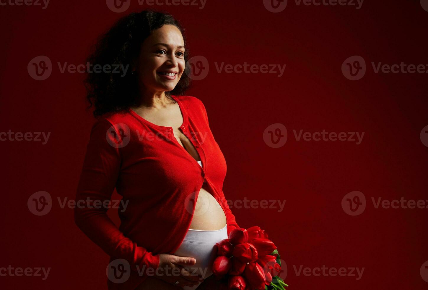 lindo feliz grávida mulher fazendo carinho dela barriga, sorridente olhando ausente, posando com uma ramalhete do tulipas, vermelho pano de fundo foto