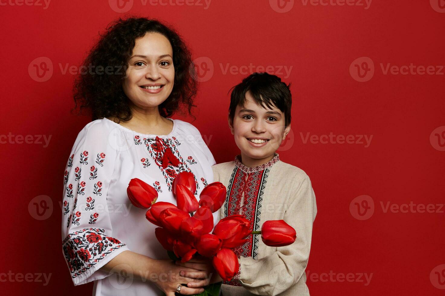 feliz sorridente étnico mulher e adolescente garoto, mãe e filho dentro tradicional ucraniano bordado camisas, segurando vermelho tulipas foto