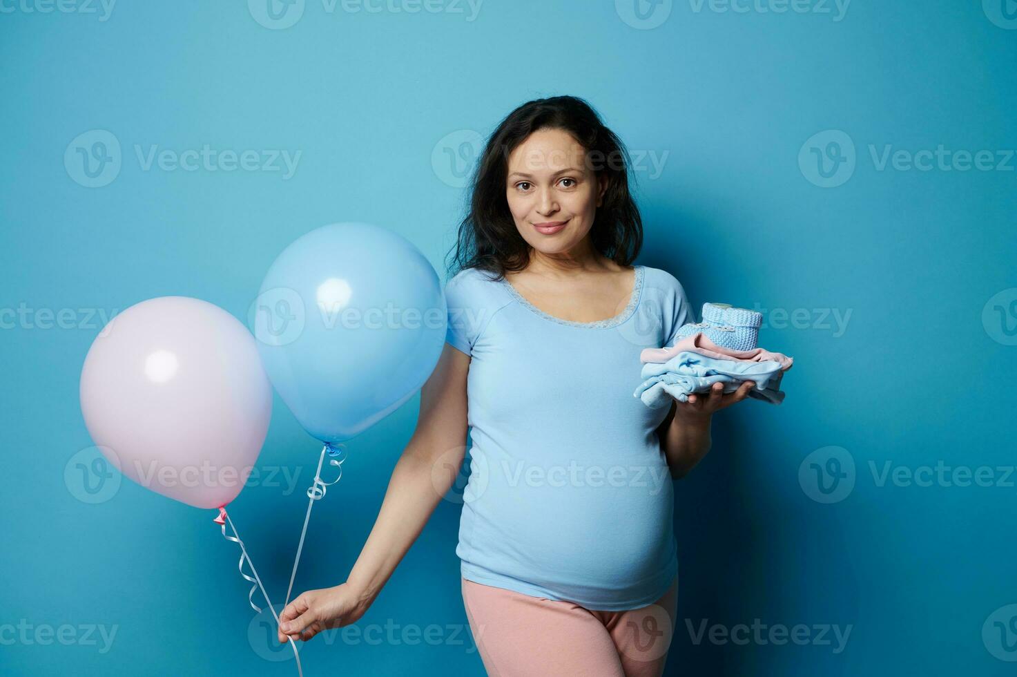 lindo grávida morena mulher posando com recém-nascido roupas e Rosa azul ar balões, isolado em azul fundo foto