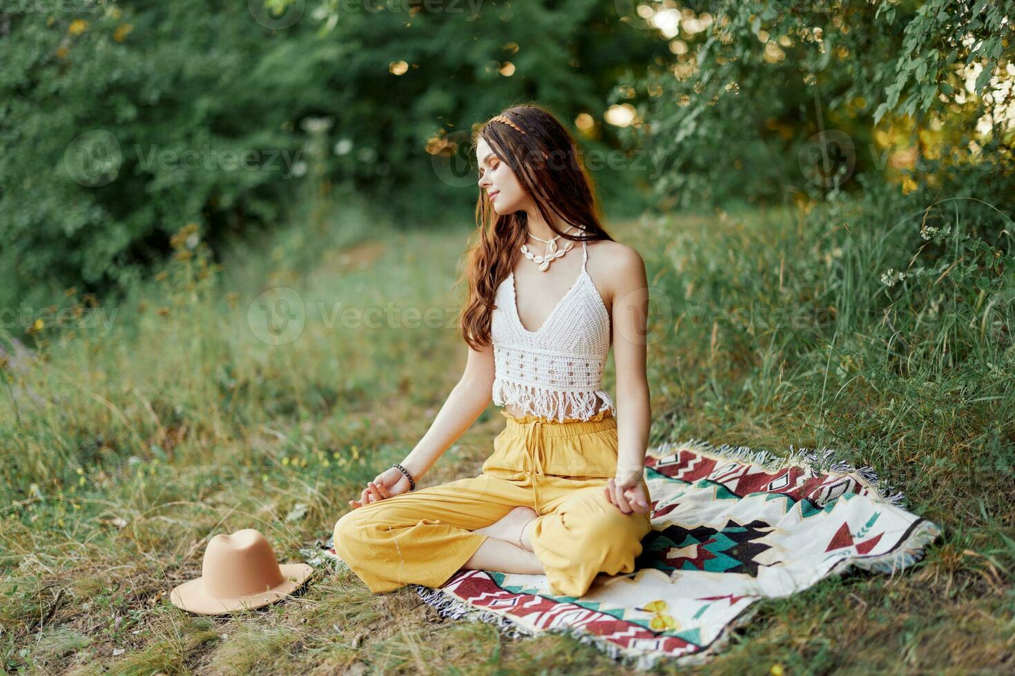uma jovem hippie mulher medita dentro natureza dentro a parque, sentado dentro uma lótus posição em dela colorida xadrez e desfrutando harmonia com a mundo dentro roupas ecológicas foto