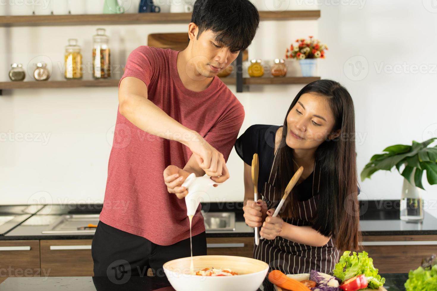 um jovem casal asiático está comendo juntos e sorrindo alegremente enquanto cozinha sua salada na cozinha. foto