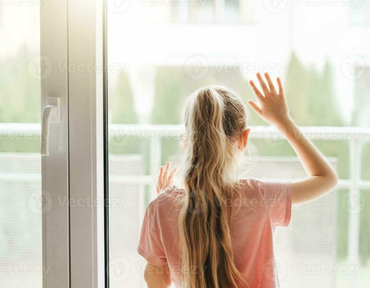 triste pequeno menina olhando através janela às lar. foto