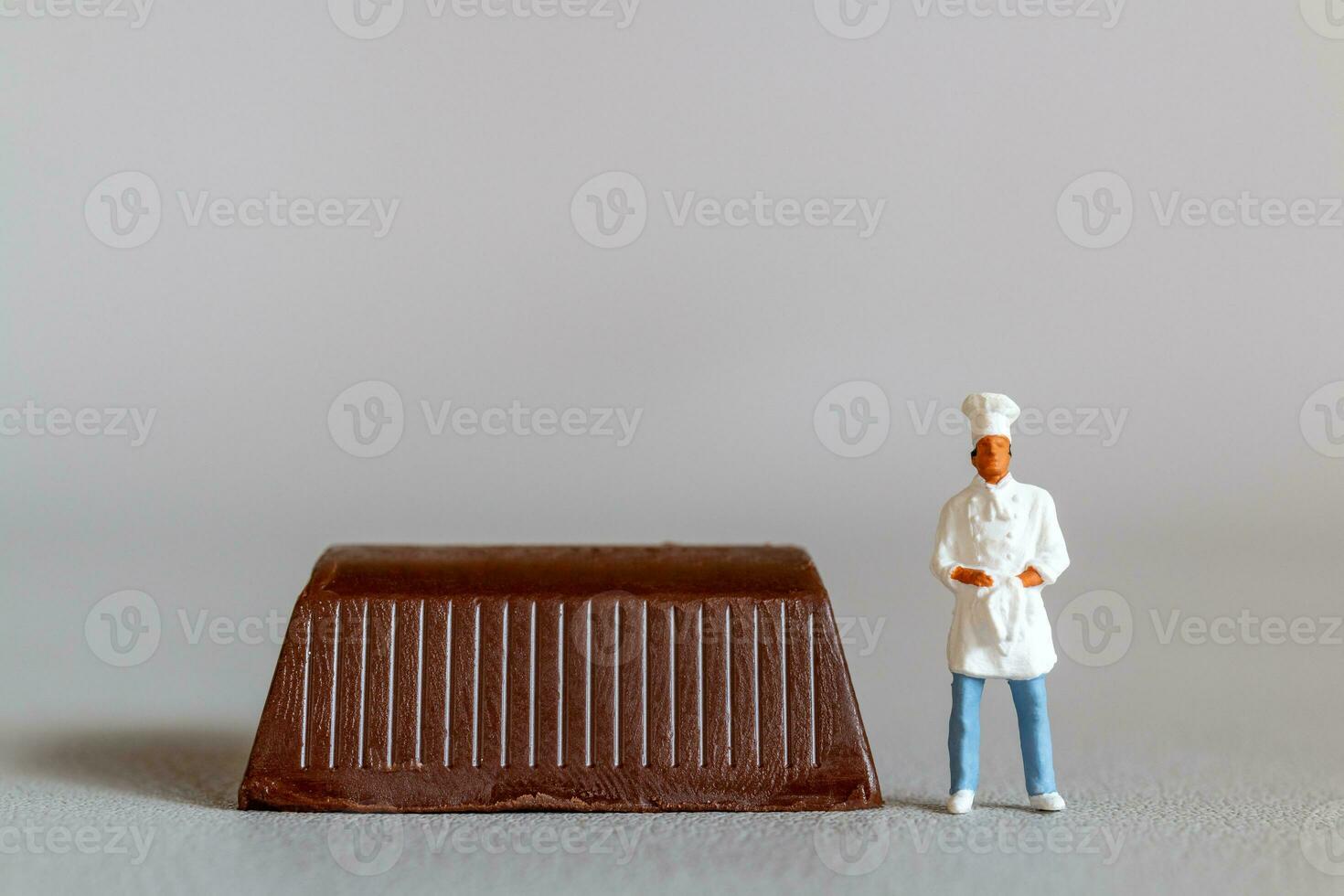 miniatura pessoas chefe de cozinha com chocolate em pé enquanto em pé contra uma cinzento fundo foto