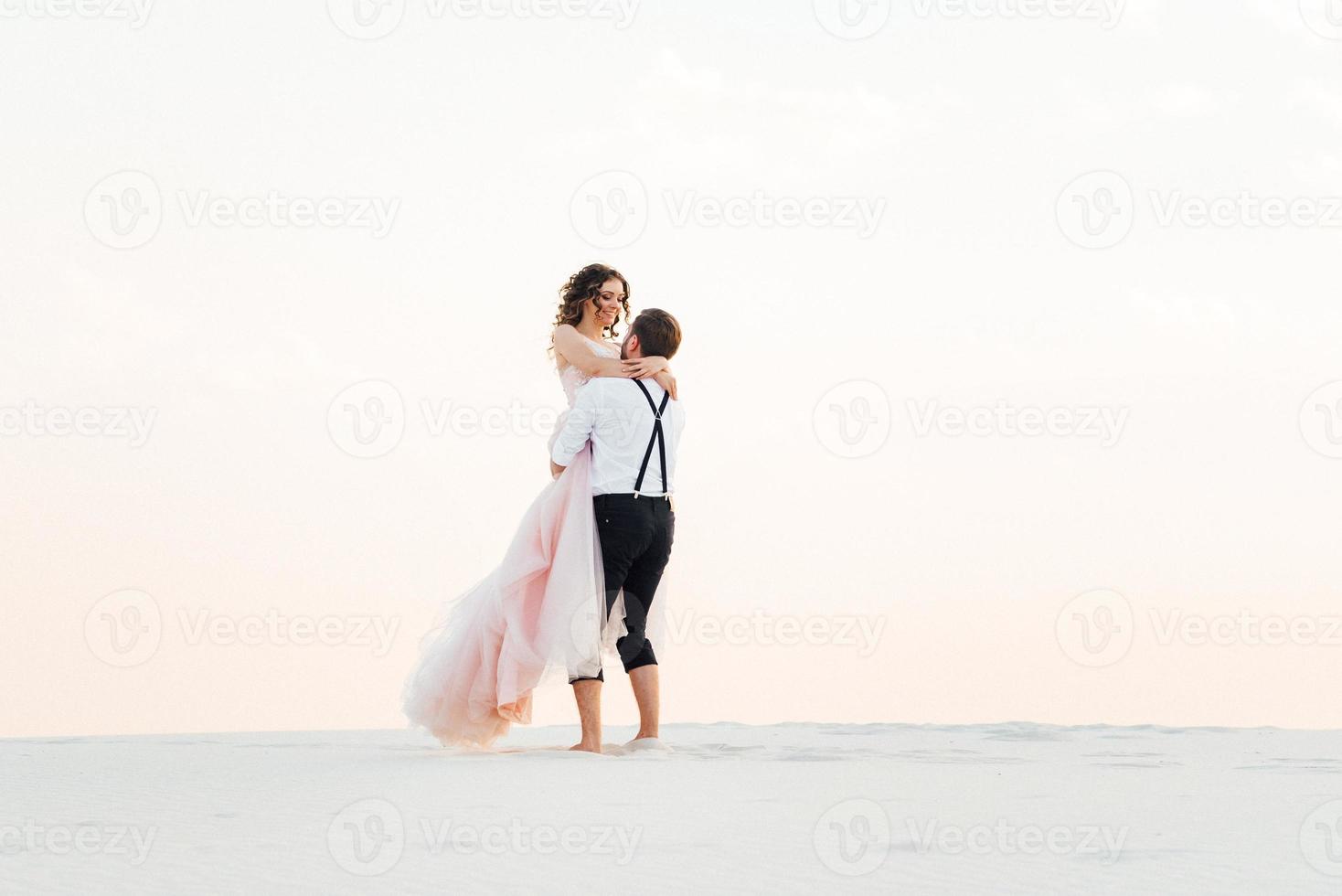 casal jovem um cara de calça preta e uma garota de vestido rosa estão caminhando na areia branca foto
