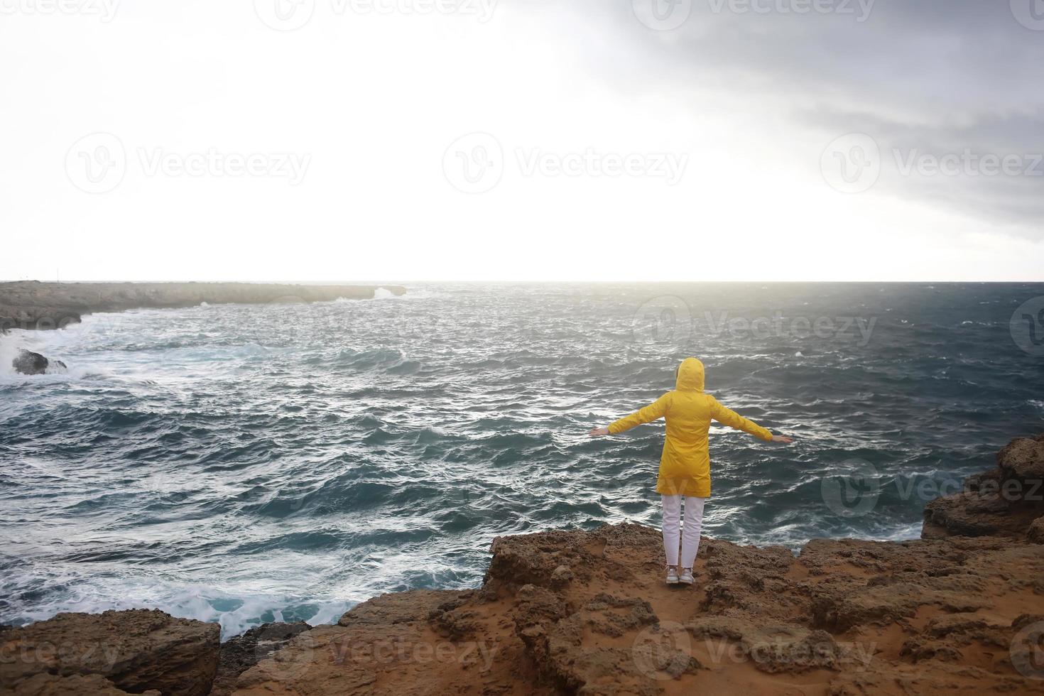 jovem vestida com uma capa de chuva amarela, de pé com os braços estendidos, enquanto aprecia a bela paisagem do mar foto