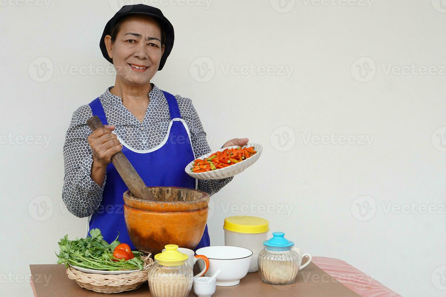 mulher sênior asiática feliz está cozinhando, usa boné e avental de chef, segura pilão, almofariz e prato de pimenta. conceito, cozinhar para a família. estilo de vida da cozinha tailandesa. atividade de idosos. foto