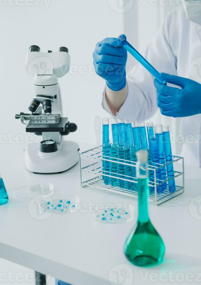 médico ou científico investigador pesquisando e experimentando multicolorido solução, frasco e microscópio dentro a laboratório ou dentro a laboratório de vestindo azul luvas e branco roupas completamente. foto