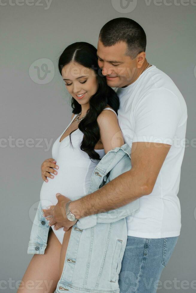 uma jovem feliz família marido e esposa estão esperando uma bebê. uma homem abraços uma grávida mulher em uma cinzento fundo foto