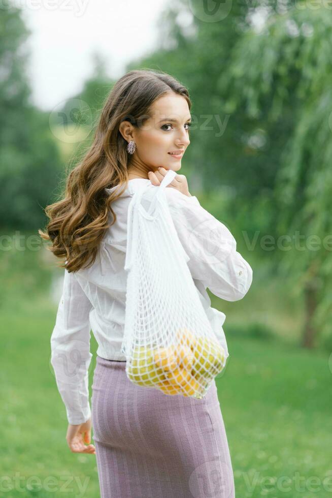 linda jovem carregando frutas com uma eco-bag de malha de algodão. andando no parque depois das compras. o conceito de lixo zero foto