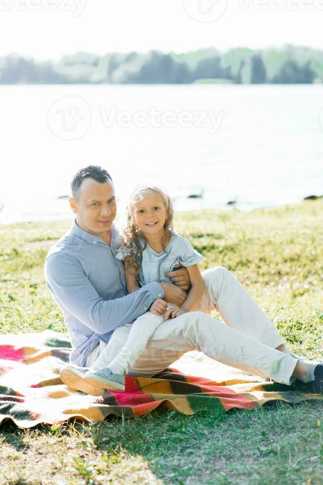 uma feliz pai detém dele filha dentro dele braços. elas ter Diversão dentro natureza, sentado em uma cobertor de a lago dentro verão foto