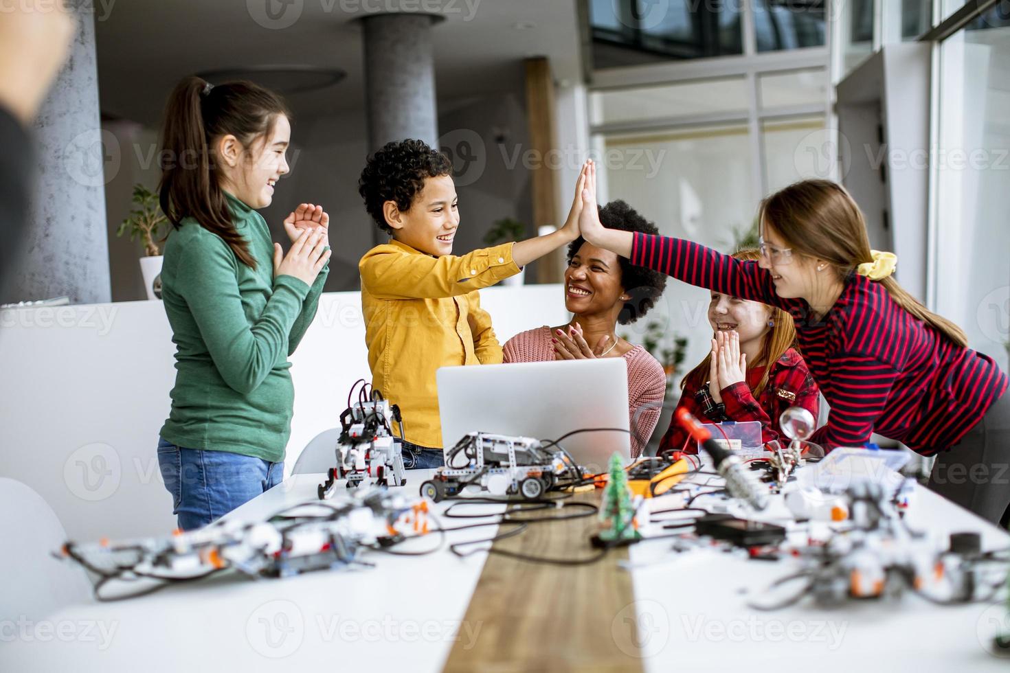 crianças felizes com sua professora de ciências afro-americana com laptop programando brinquedos elétricos e robôs na sala de aula de robótica foto