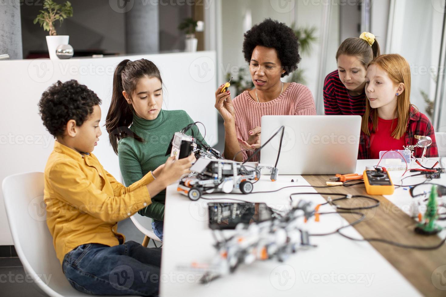 crianças felizes com sua professora de ciências afro-americana com laptop programando brinquedos elétricos e robôs na sala de aula de robótica foto